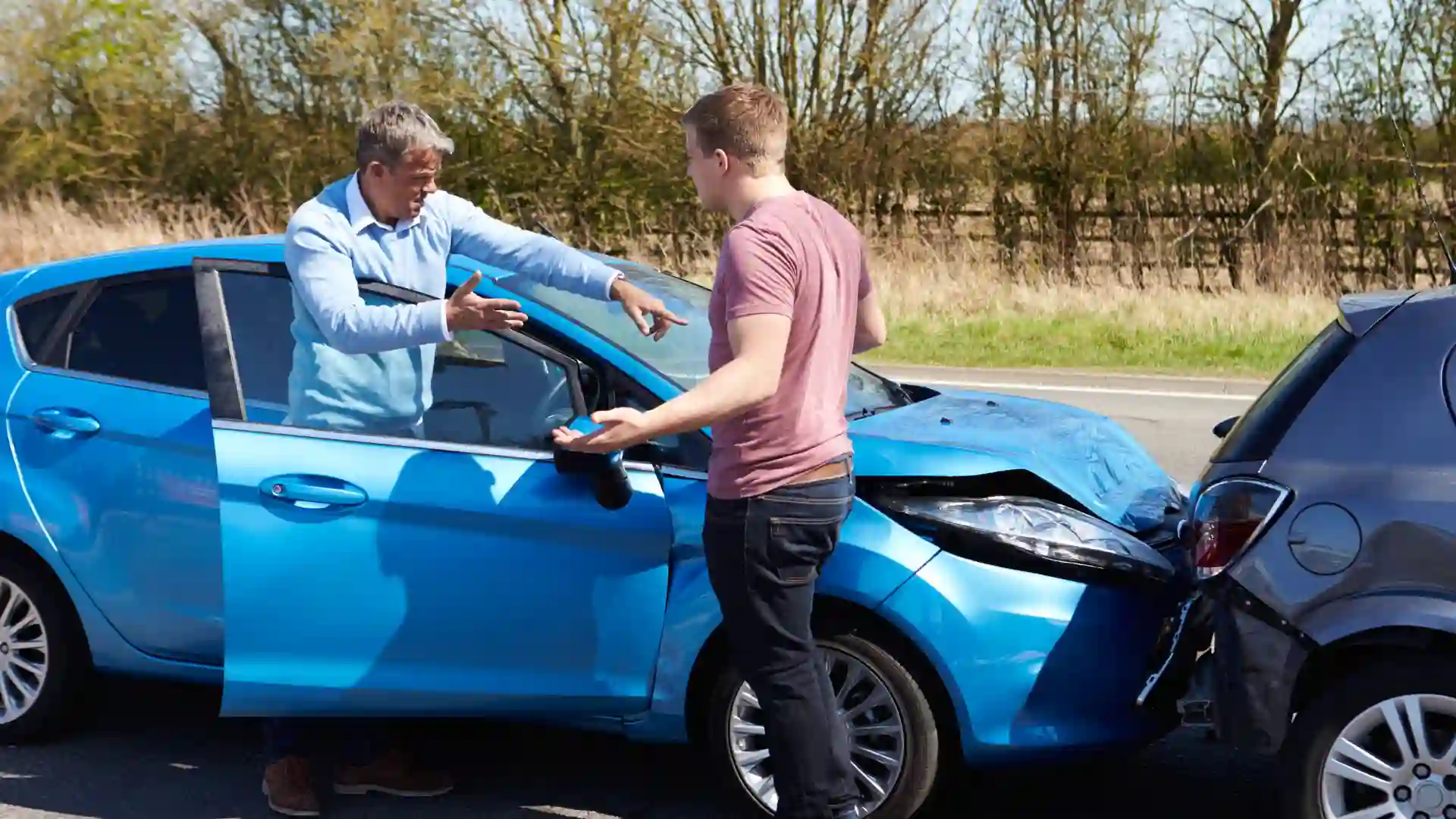Two men arguing beside a damaged blue car after an accident.