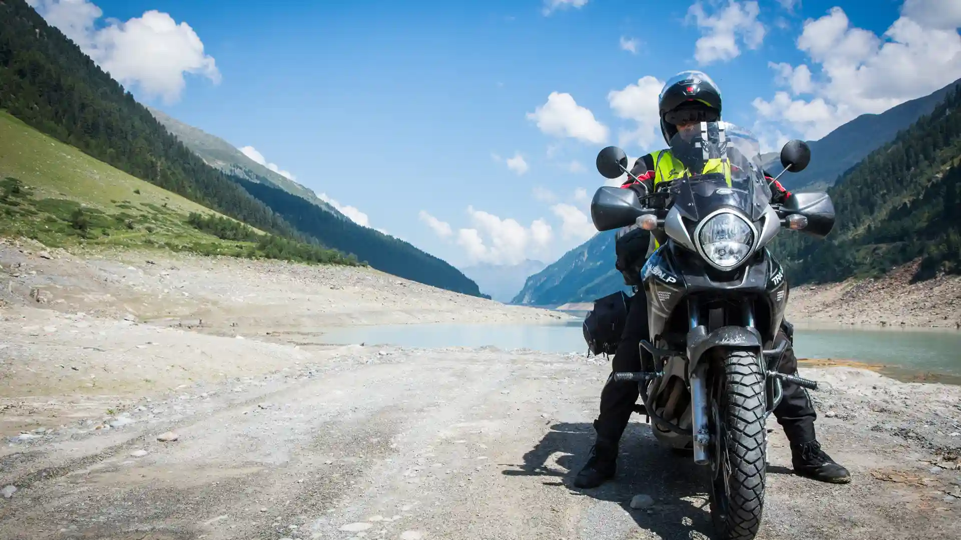 Motorcyclist in protective gear standing by a bike on a scenic mountain road.