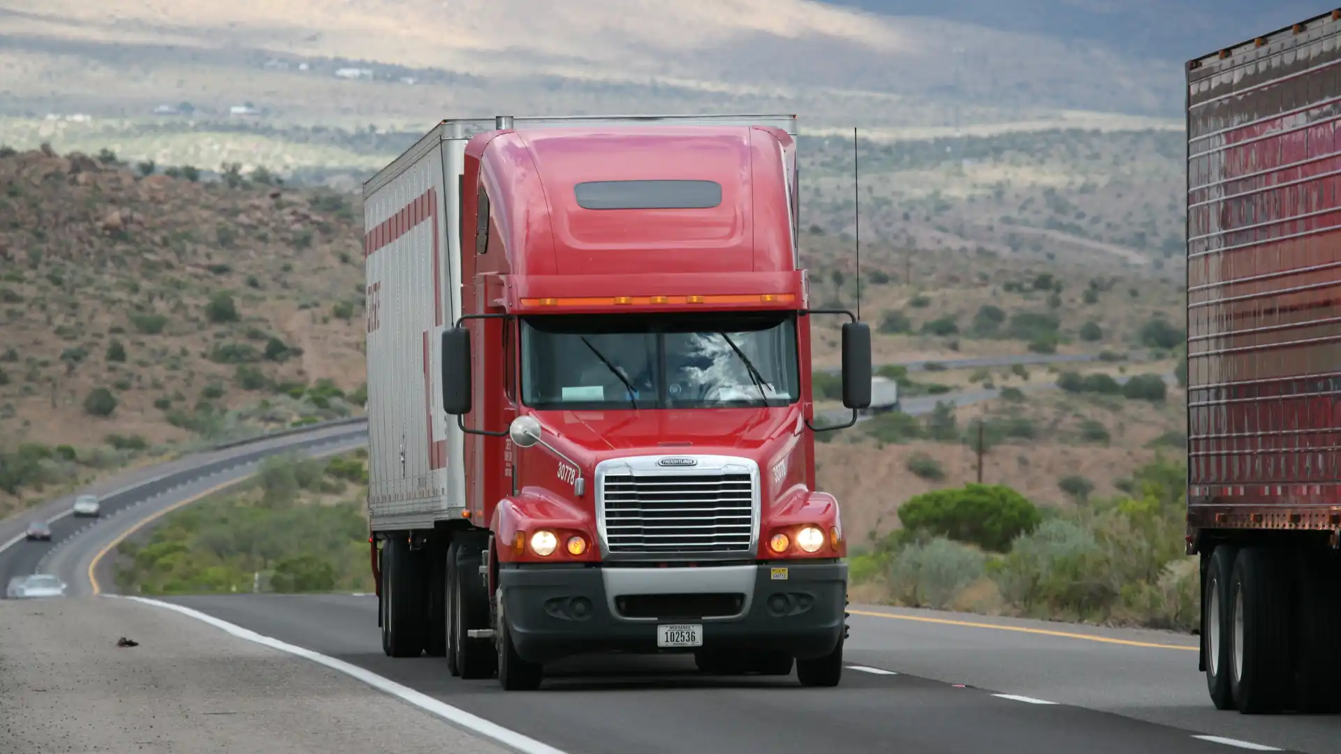 Red semi-truck driving on a highway with mountains in the background."